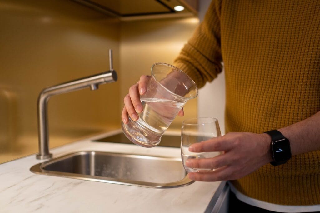 A kitchen faucet with water flowing into a clear glass, but the water has a slight brownish tint, indicating impurities. The background shows a typical kitchen setting with neutral tones.