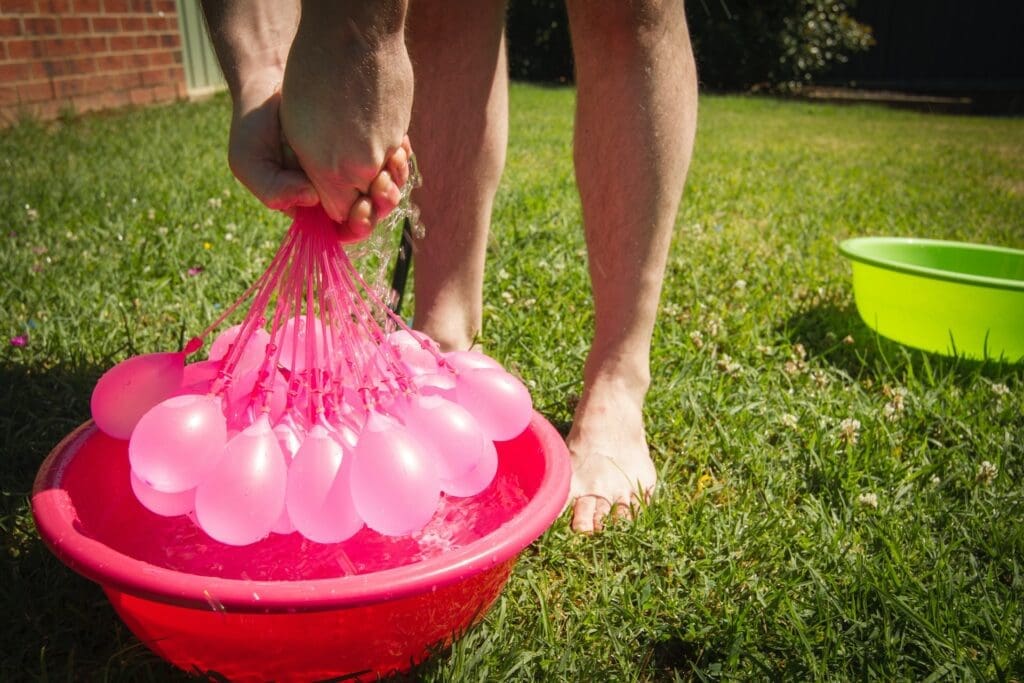A backyard scene with a family gathered around a table, enjoying a barbecue. A child fills a water balloon from an outdoor faucet, with the water looking crystal clear.