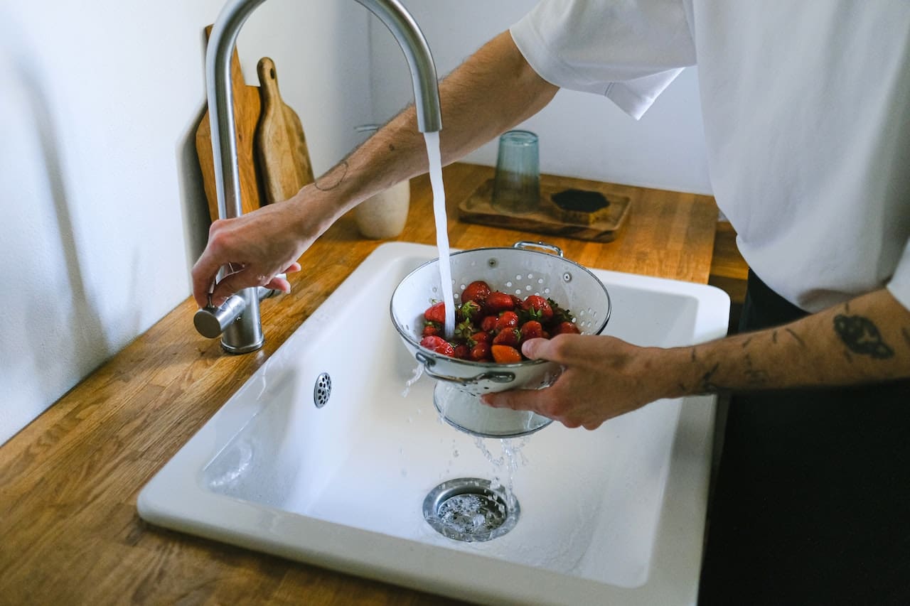 An indoor scene of a modern kitchen with large windows. A woman is rinsing fresh fruits under a stream of clear water in the sink, while a child drinks from a glass filled with sparkling water.