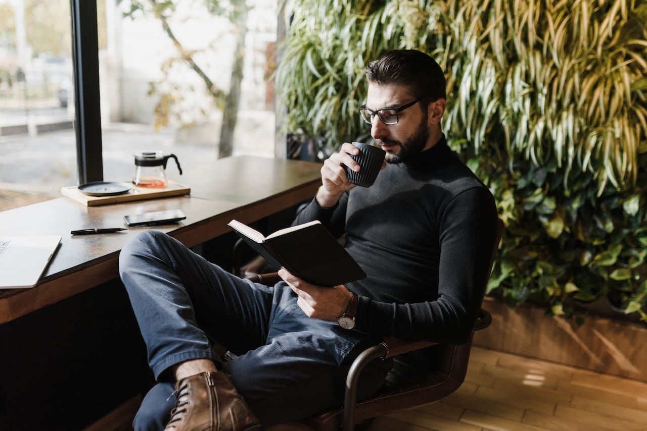 A cozy living room scene with large windows allowing sunlight to filter in. A man relaxes on a couch, reading a book, with a glass pitcher of clear water and a filled glass on a coffee table next to him.