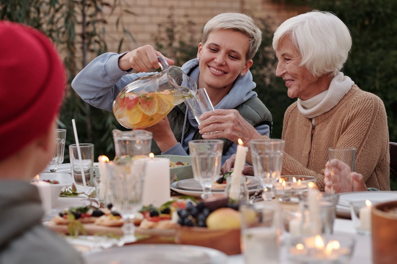 A spacious patio area with a family gathered around a table, sharing a meal. A large glass jug of iced water, garnished with cucumber slices and rosemary sprigs, sits in the center of the table. In the background, a garden fountain trickles water soothingly.