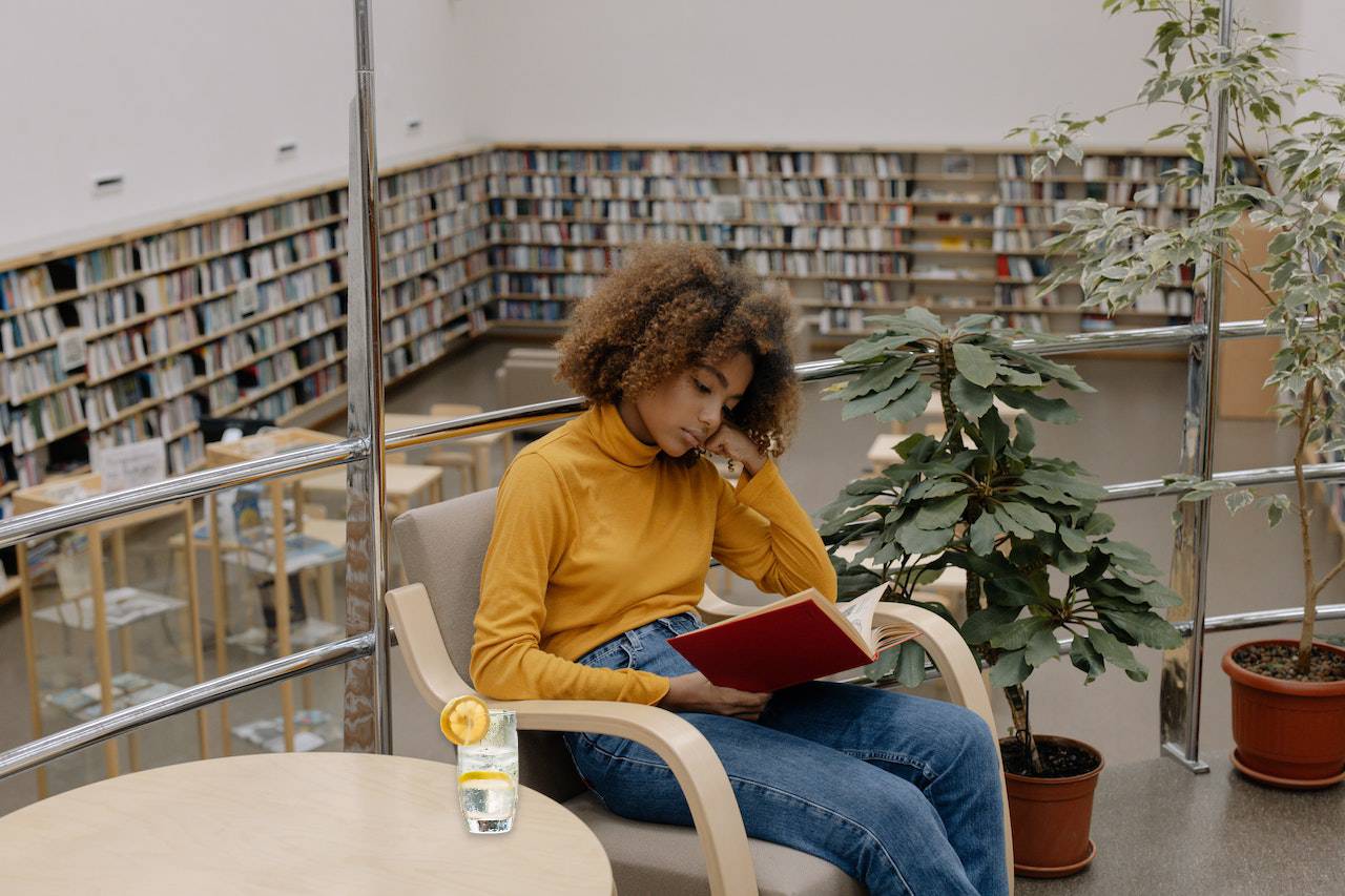 A study room with bookshelves lining the walls. A scholar reads, with a glass pitcher of water adorned with rosemary sprigs and lemon slices on the desk.