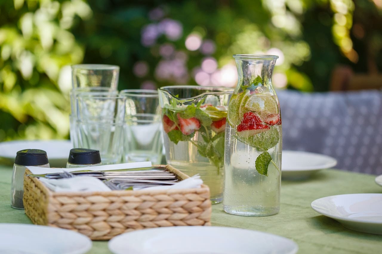 An open-concept kitchen and dining area illuminated by natural light. A family gathers around the dining table, sharing a meal. The centerpiece is a large glass pitcher filled with ice-cold water, lemon slices, and fresh mint leaves.