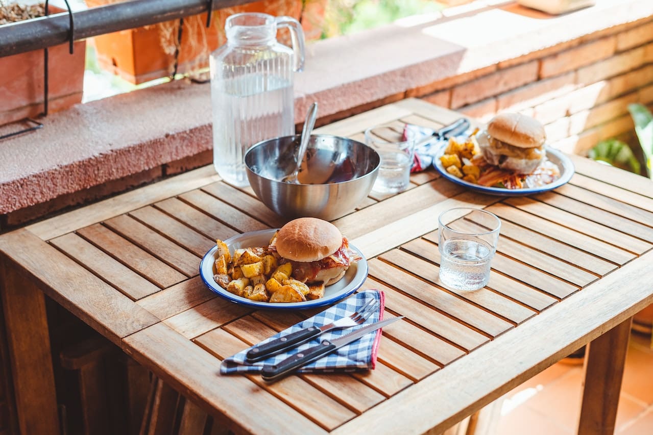 A rustic kitchen with wooden beams and large windows. A family gathers around a wooden table, sharing a meal, with a large glass jug of iced water in the center.