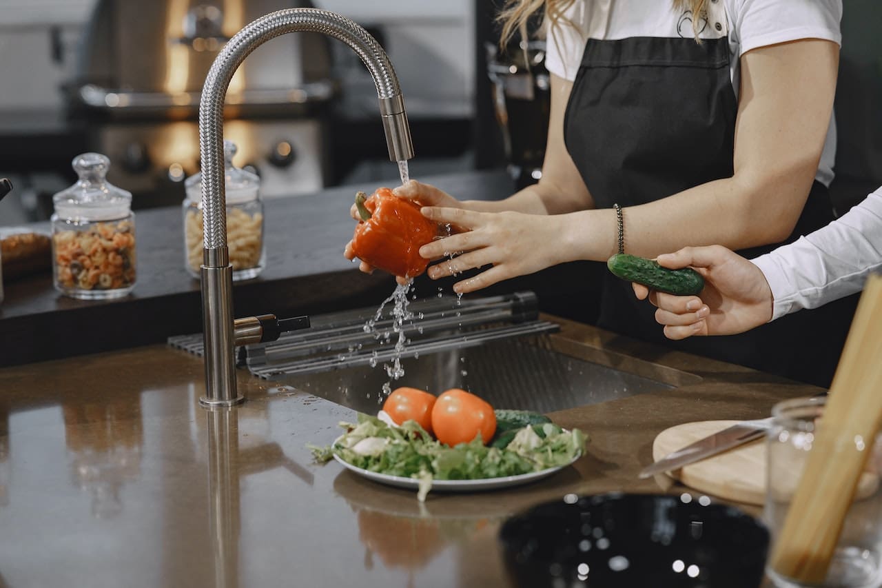 A picture of a homemaker washing vegetables in slighlty discolored tap water with children watching over in convern caused by unfiltered water, highlighting the need for clean water for daily use in Laredo households.