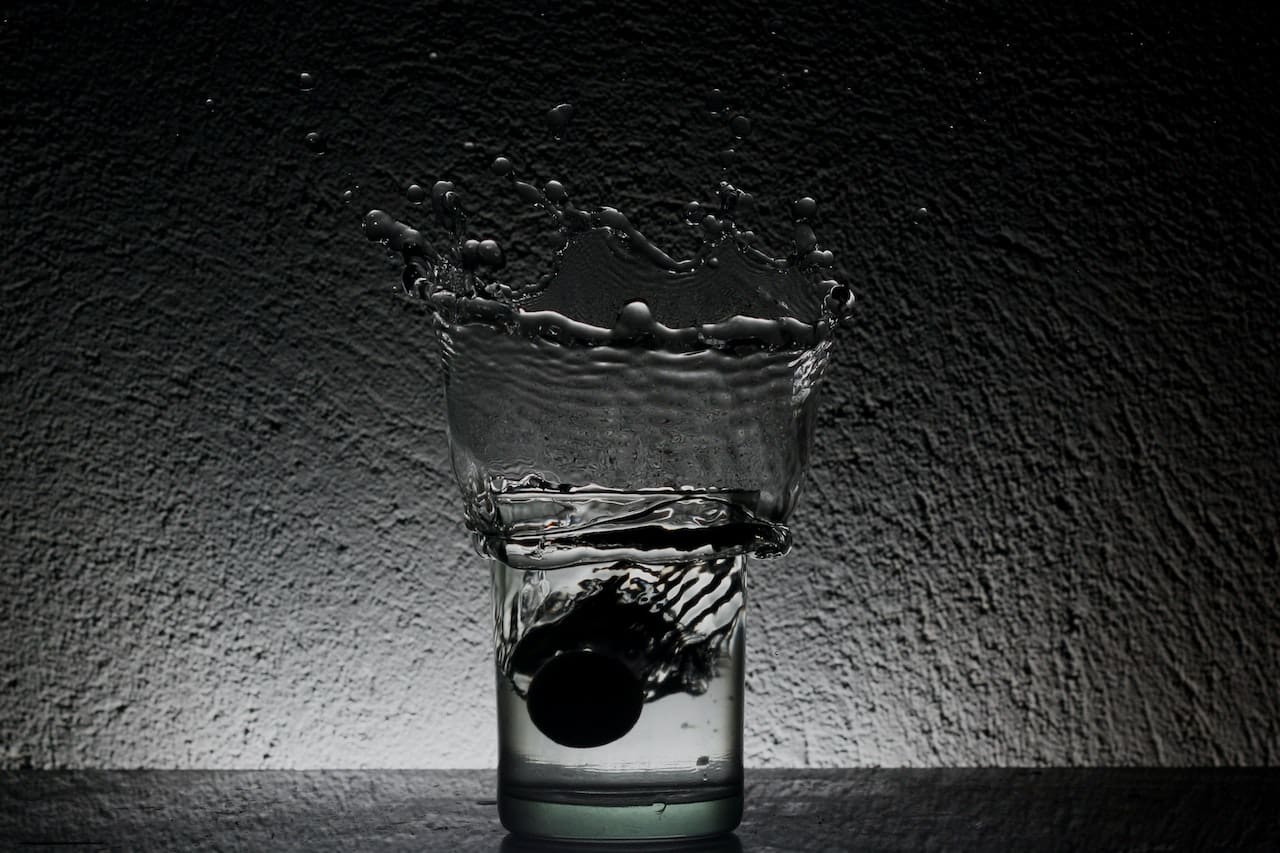 A close-up of a clear glass on a kitchen countertop, filled with water that has a slight brownish tint. Nearby, a dropper dispenses a single drop into the glass.