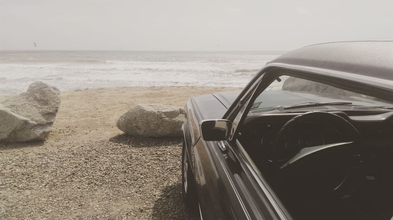 A close-up of a car windshield covered in water spots and streaks. The blurred background suggests the car is parked near a beach, with palm trees and the ocean faintly visible.