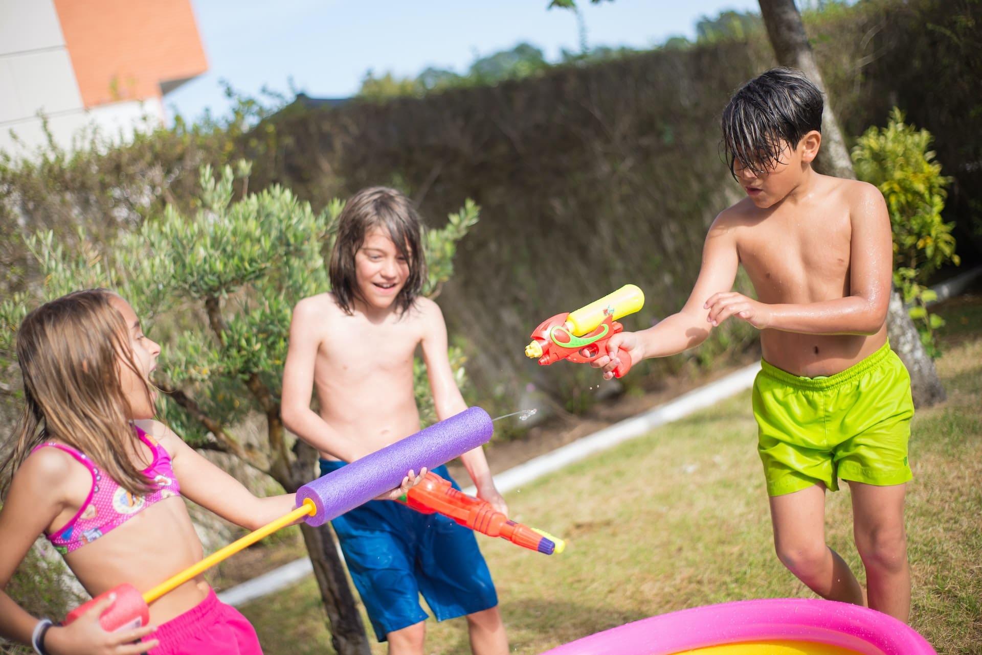 A family backyard barbecue scene. Children play with water guns, adults chat around a table with a pitcher of iced water garnished with lemon slices, and a sprinkler system ensures the lawn stays lush.