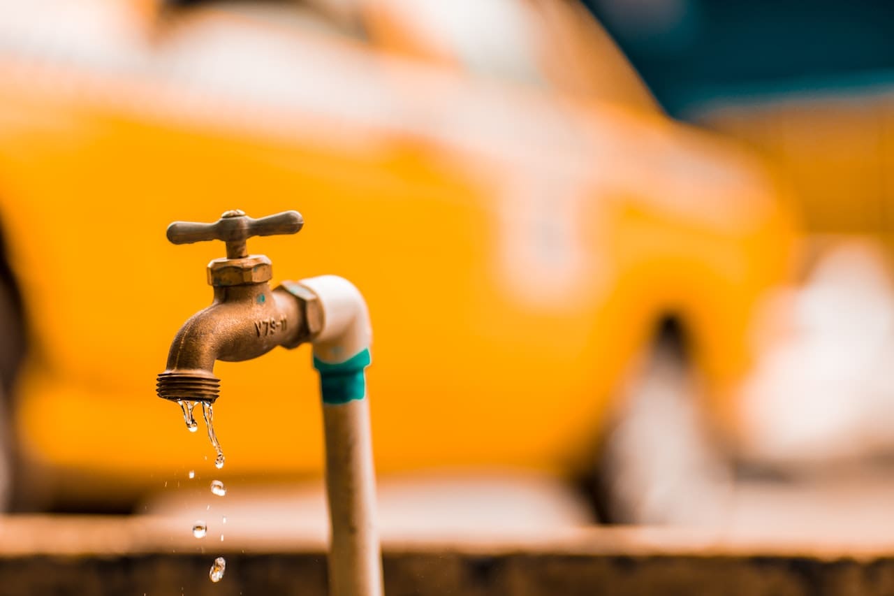 A close-up of a dripping outdoor faucet attached to a brick wall. The droplet about to fall has a magnified image of a bacteria or virus within.