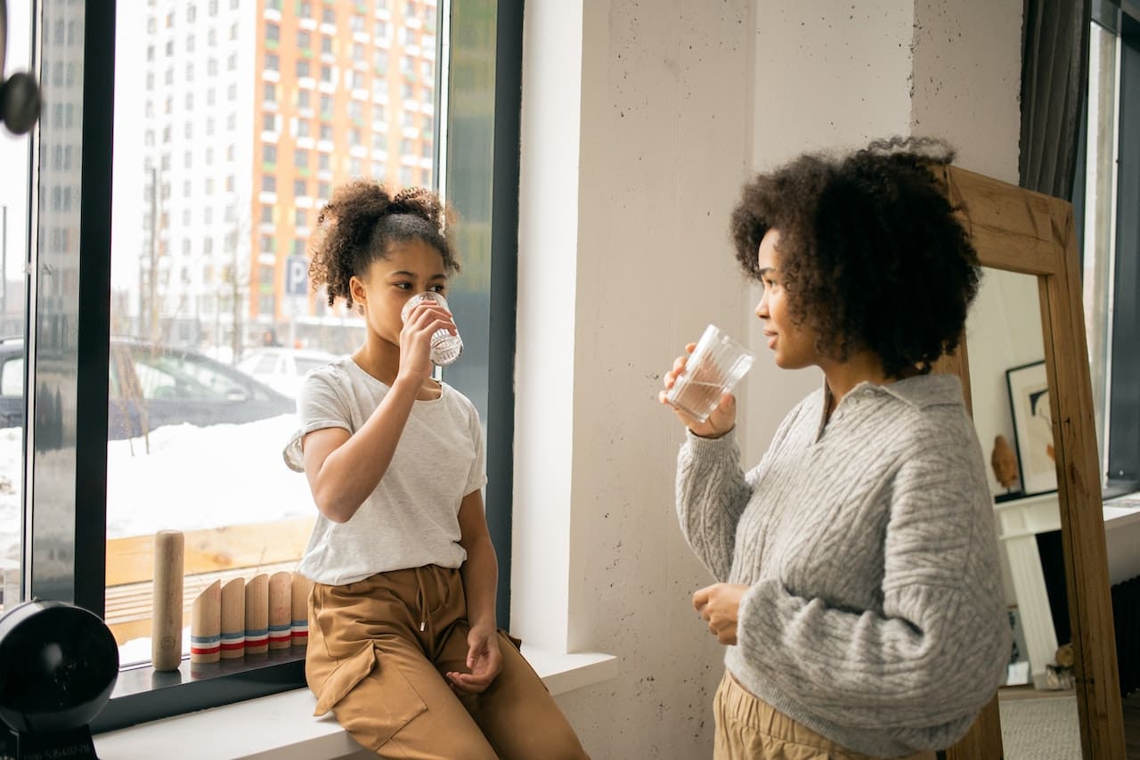 A sunlit kitchen scene with a large window overlooking a verdant backyard. A mother and child stand by the kitchen island, filling up reusable water bottles from a sleek, modern faucet, with the water appearing crystal clear.