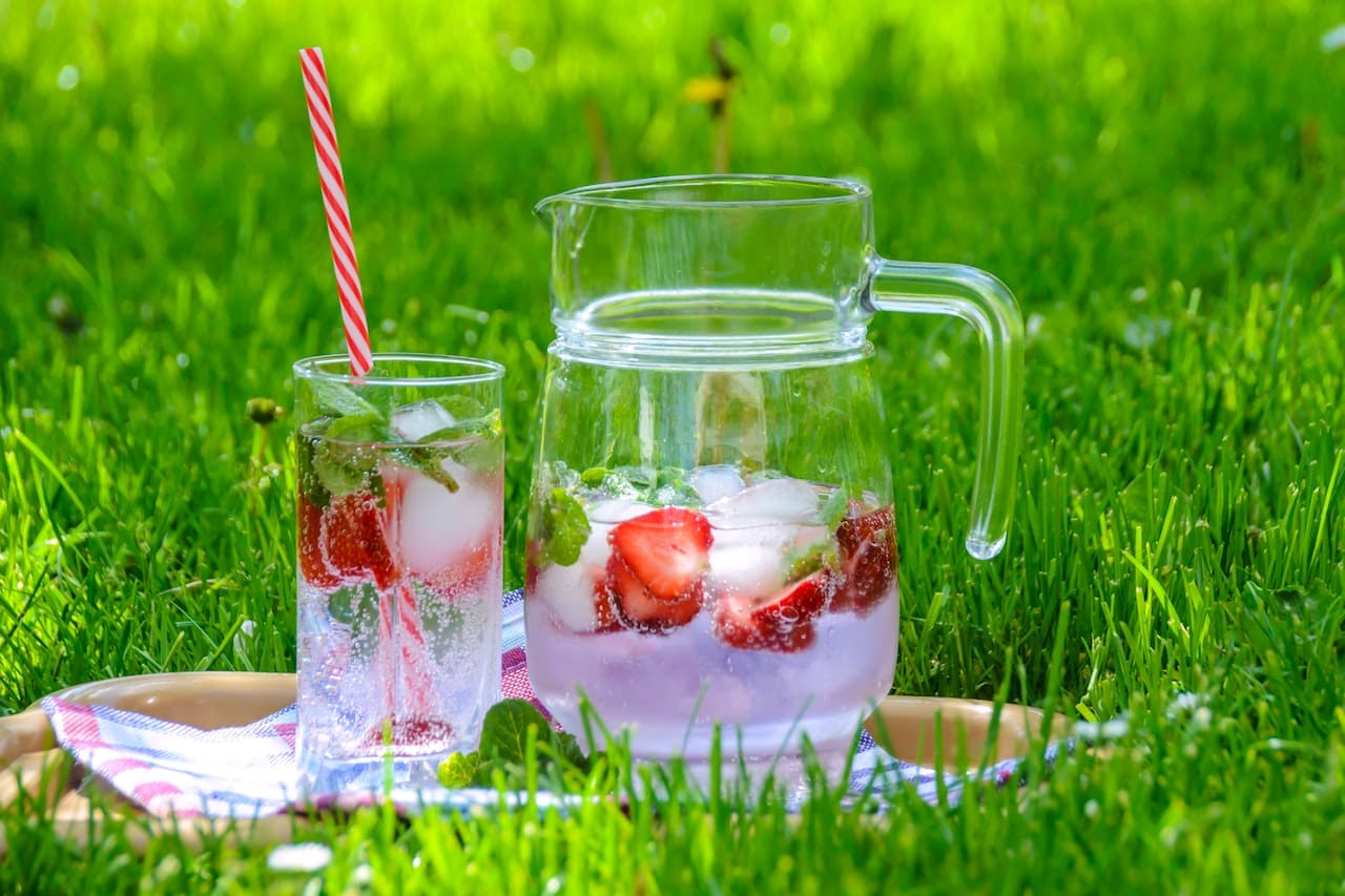 A backyard scene with children laughing and playing under a sprinkler. Nearby, a table is set for a meal, with a pitcher of clear water adorned with fresh mint and lemon slices.