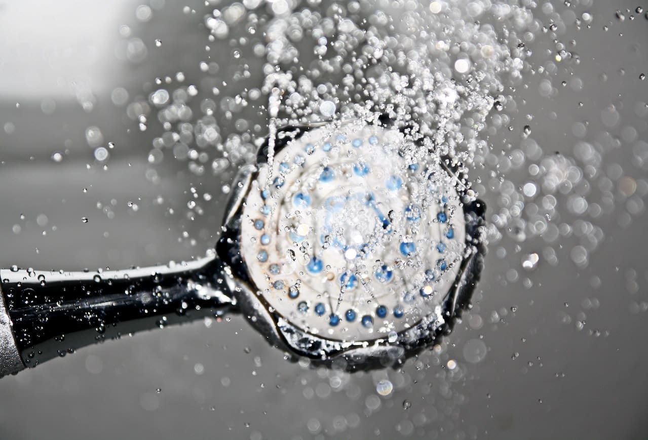 A close-up of a showerhead in a bathroom, with water droplets suspended mid-spray. The showerhead's surface has noticeable white calcium deposits, indicating hard water.