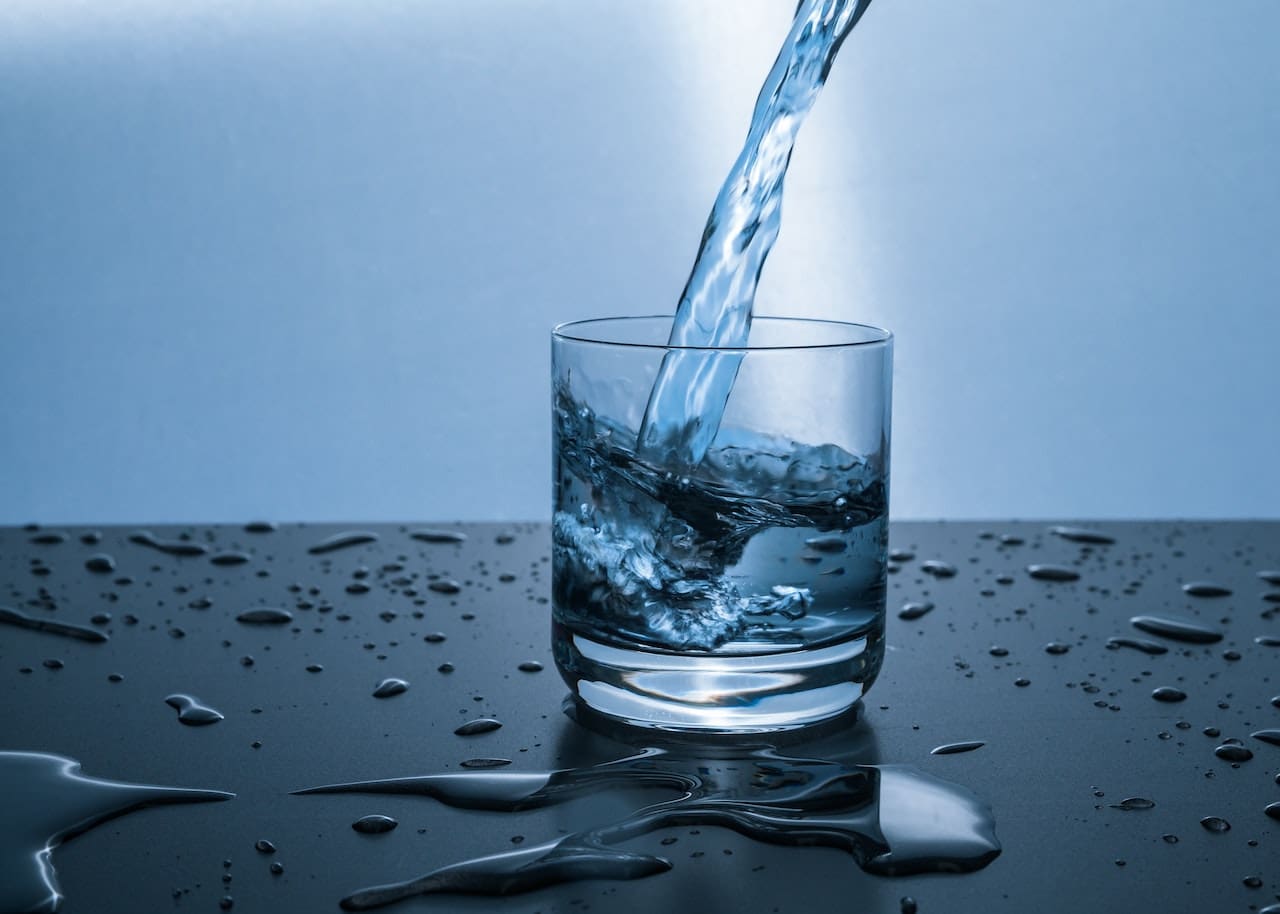 A close-up of a glass on a kitchen counter filled with water. Floating on the surface are tiny particles, and the water has a cloudy appearance, indicating impurities.