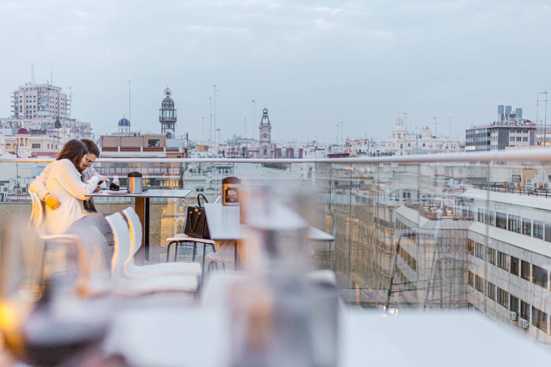 A rooftop terrace of a high-rise building, overlooking the city and bay. Residents lounge, enjoying the view, with a sleek water dispenser offering chilled, filtered water to guests.